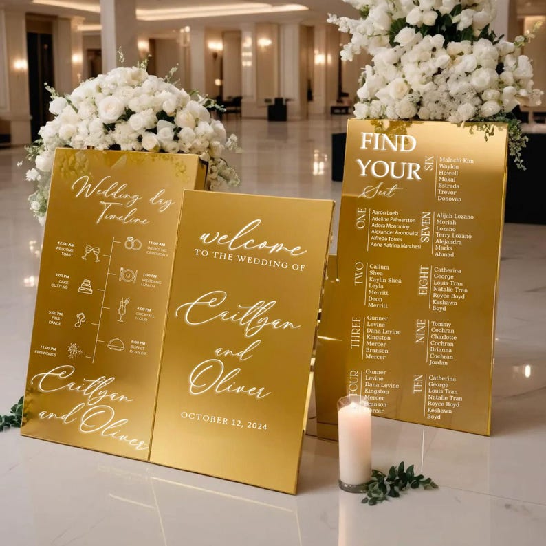 May include: Gold mirror acrylic wedding signs with white calligraphy. The signs include a wedding timeline, welcome sign, and seating chart. The seating chart is labeled "Find Your Seat" and lists guest names by table number.