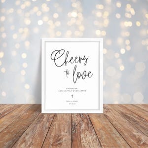 May include: White framed sign with black calligraphy that reads "Cheers to love, Laughter, and happily ever after. Cora + Henry 07.15.XX".