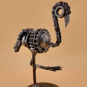 Puede incluir: Una escultura de metal de un flamenco hecha con materiales reciclados, como cadenas y engranajes. La escultura está pintada en un acabado metálico y se apoya sobre una base redonda.