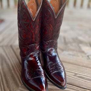 May include: A pair of dark cherry red leather cowboy boots featuring a pointed toe, a stacked heel, and intricate tonal stitching patterns across the shaft and vamp. The boots have a polished finish and a tan leather lining visible at the top.