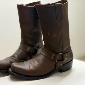 Puede incluir: Un par de botas de cuero marrones para motocicleta. Las botas tienen una puntera cuadrada, una correa de arnés con hebilla metálica y caña alta. El cuero tiene un aspecto desgastado con algunas rozaduras.