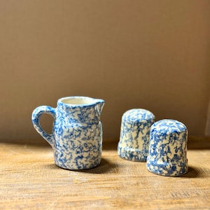 May include: A ceramic creamer pitcher with a handle and two matching shakers, all featuring a white base with a blue speckled sponge-painted pattern. The set consists of three individual pieces with a consistent mottled blue and white finish.