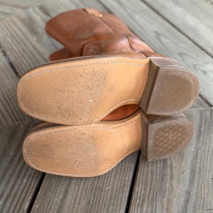 Pu&ograve; includere: Un paio di stivali da cowboy in pelle color cuoio con punta quadrata, cuciture visibili lungo i bordi delle suole chiare e tacchi in legno a strati. Gli stivali sono posizionati in modo da mostrare le suole, dalla consistenza liscia e usurata.