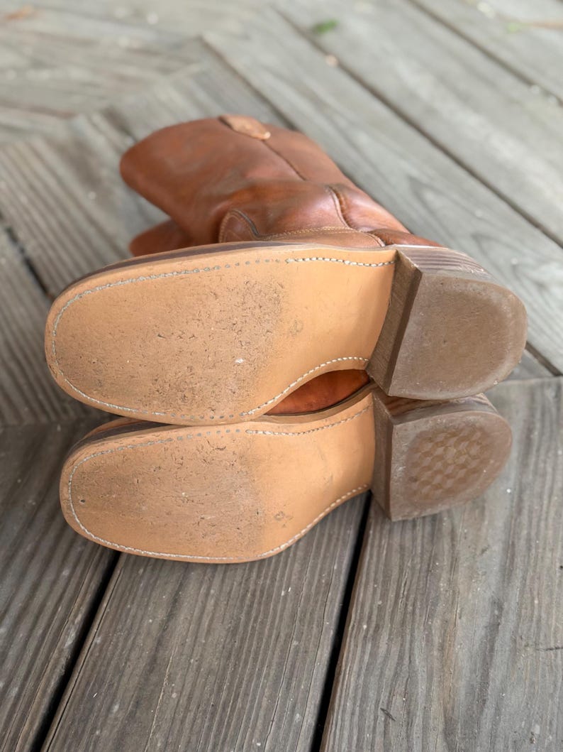 Pu&ograve; includere: Un paio di stivali da cowboy in pelle color cuoio con punta quadrata, cuciture visibili lungo i bordi delle suole chiare e tacchi in legno a strati. Gli stivali sono posizionati in modo da mostrare le suole, dalla consistenza liscia e usurata.