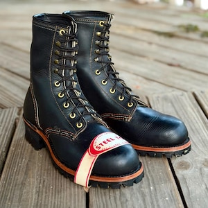 Puede incluir: Un par de botas de trabajo de cuero negro con puntera de acero, ojales metálicos dorados, cordones negros y suela de goma gruesa con tacos. Una etiqueta blanca y roja está unida a la parte delantera con el texto STEEL TOE claramente visible.