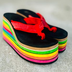 May include: A pair of platform flip-flops with a rainbow-striped sole and black footbed. The straps are red. The platform sole is approximately 3 inches high.