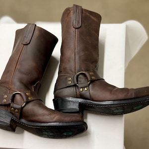 以下が含まれることがあります： 茶色の革製バイクブーツのペア。ブーツは、真鍮色の金具が付いたハーネスストラップ、スクエアトゥ、スタックヒールが特徴です。革は使い古された外観をしています。