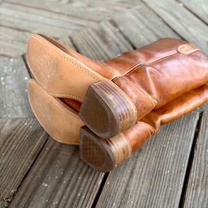 Pu&ograve; includere: Un paio di stivali da cowboy in pelle color cuoio con finitura liscia, gambale a met&agrave; polpaccio e tacco in legno a strati. Le suole sono in pelle chiara con cuciture visibili lungo i bordi, mostrando una consistenza usurata sul fondo.