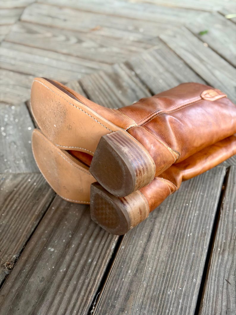 Pu&ograve; includere: Un paio di stivali da cowboy in pelle color cuoio con finitura liscia, gambale a met&agrave; polpaccio e tacco in legno a strati. Le suole sono in pelle chiara con cuciture visibili lungo i bordi, mostrando una consistenza usurata sul fondo.