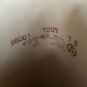 Pu&ograve; includere: L'interno di una scarpa con testo stampato che include 98001, 443, 7207, 468, 7 E, MADE IN U.S.A., INNERSOLE FORTIFIED FIBRE, LINING MAN MADE MATERIAL e un logo circolare con la lettera A. Lo sfondo &egrave; di colore crema chiaro.
