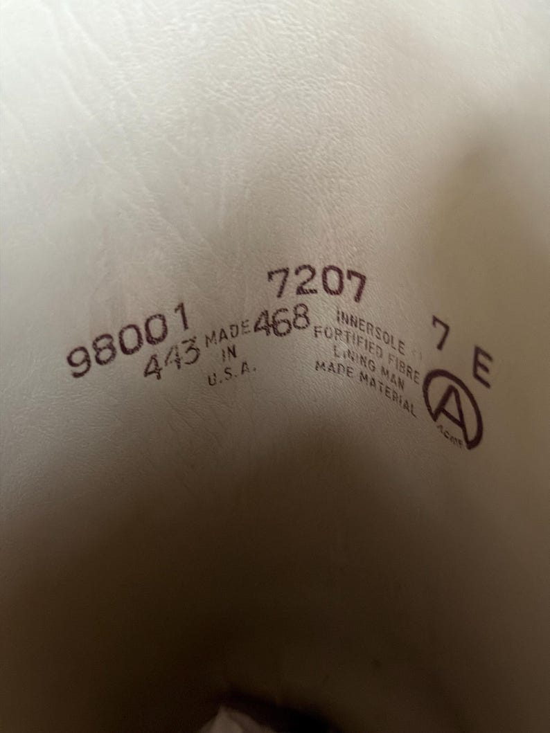 Pu&ograve; includere: L'interno di una scarpa con testo stampato che include 98001, 443, 7207, 468, 7 E, MADE IN U.S.A., INNERSOLE FORTIFIED FIBRE, LINING MAN MADE MATERIAL e un logo circolare con la lettera A. Lo sfondo &egrave; di colore crema chiaro.