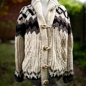 Op de afbeelding: Een beige kabelgebreid vest met een sjaalkraag en houten knevelsluitingen. De trui heeft een geometrisch patroon in zwart en donkergroen over de borst en schouders. Het kledingstuk wordt op een mannequin getoond.