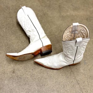 May include: A pair of white cowboy boots with pointed toes. The boots feature decorative stitching and black trim. The heels are brown and the soles show wear. The boots are angled on a concrete surface.