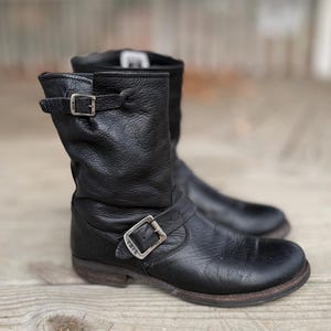 Könnte beinhalten: Ein Paar schwarze Leder-Ankle Boots mit silberfarbenen Schnallen. Die Stiefel haben ein abgenutztes Aussehen und einen kurzen Schaft. Der Markenname Frye ist auf einer der Schnallen sichtbar. Die Stiefel werden auf einer Holzoberfläche präsentiert.