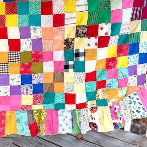 May include: A patchwork quilt constructed from numerous small square fabric pieces in a wide variety of colours and patterns, including solid red, yellow, green, blue, and pink, alongside floral, plaid, and polka dot prints, finished with a ruffled edge.