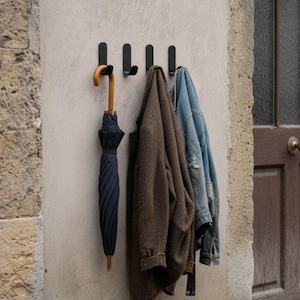 Pode incluir: Um cabide de parede com quatro ganchos pretos. Um guarda-chuva azul marinho com cabo de madeira está pendurado no primeiro gancho. Um casaco castanho e uma jaqueta jeans azul estão pendurados nos outros ganchos.