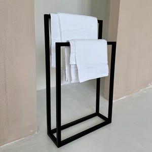 Pode incluir: Um toalheiro de metal preto com dois níveis, segurando toalhas brancas dobradas. O suporte tem um design de estrutura retangular, adequado para um banheiro ou spa. As toalhas parecem macias e fofas.