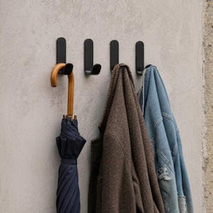 Può includere: Un attaccapanni a parete con quattro ganci neri. Un ombrello blu navy con manico in legno è appeso al primo gancio. Un cappotto marrone e una giacca di jeans blu sono appesi agli altri ganci.