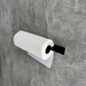 May include: A white paper towel roll is mounted on a black metal holder against a gray concrete wall. The paper towel roll is partially unrolled, with a single sheet hanging down. The holder is a simple, modern design.