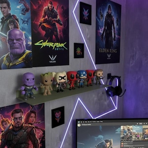 Puede incluir: Una estantería verde exhibe figuras coleccionables, incluyendo personajes de Marvel y otras franquicias. La pared muestra carteles de videojuegos como Cyberpunk 2077 y Elden Ring, junto con arte enmarcados y auriculares.