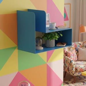 May include: A modern, blue wall-mounted shelf unit with two levels. The top shelf holds a framed photo, a white mug, and a coaster. The lower shelf displays a potted plant, a glass cloche, and a small notebook. The background features a colorful geometric pattern.