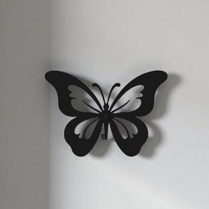 May include: A black metal butterfly wall decor. The butterfly has a symmetrical design with cut-out details in the wings. It is mounted on a white wall, creating a contrast. The butterfly is a decorative item.