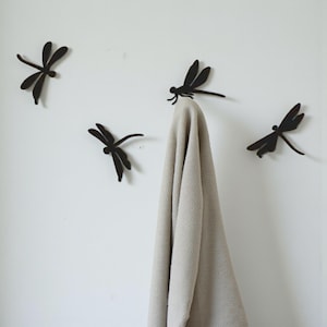 May include: Four black metal dragonfly wall hooks are mounted on a white wall. One hook holds a beige towel. The dragonfly hooks have detailed wings and bodies, adding a decorative touch to any space. The hooks are spaced apart.