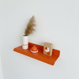 Könnte beinhalten: Ein orangefarbenes Metallregal mit einer weißen Vase, einer kleinen orangefarbenen Kerze und einem Holzblock. Die Vase hat eine braune Pflanze und ein weißes und orangefarbenes Design. Die Kerze steht auf einem weißen Teller. Der Holzblock hat ein weißes und orangefarbenes Design.