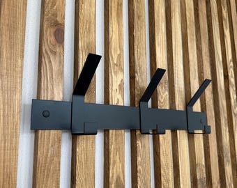 May include: A black metal wall-mounted coat rack with four hooks. The rack is mounted on a vertical wooden slat wall with varying shades of brown. The hooks are designed for hanging coats, hats, or other items.