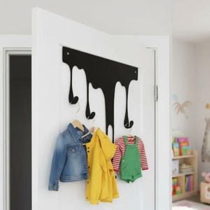 May include: A black metal wall-mounted coat rack with a unique dripping design. Three hooks hold a blue denim jacket, a yellow raincoat, and green overalls with a red and white striped shirt. The rack is mounted on a white door.