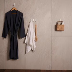 May include: A dark grey robe hangs on a wooden hanger. A white towel and a wooden back brush hang on hooks. A small woven basket holds rolled towels and a container, against a neutral tiled wall.