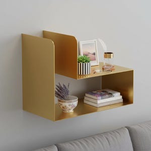 May include: A gold-colored, two-tiered wall shelf with rounded edges. The top shelf holds a small succulent in a striped pot, a framed picture, and a glass with a feather. The bottom shelf displays books and a decorative bowl.
