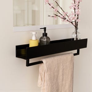 May include: A black metal bathroom shelf with a towel rail, holding a yellow soap dispenser, a dark glass soap dispenser, and a clear glass vase with pink flowers. A beige towel hangs from the rail. The shelf is mounted on a white wall.