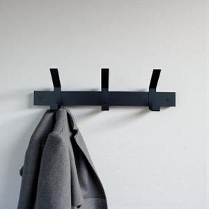 Puede incluir: Un perchero de pared azul oscuro con tres ganchos. Un abrigo gris cuelga de uno de los ganchos. El perchero está montado en una pared blanca.