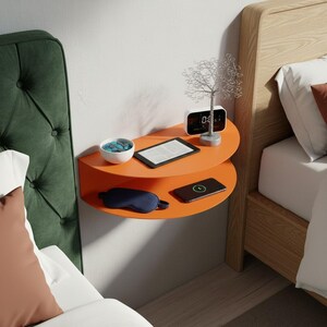 Peut inclure: Une table de chevet murale orange semi-circulaire à deux étagères. L'étagère supérieure contient un petit bol blanc avec des pierres bleues, une tablette et un réveil numérique. L'étagère inférieure a un masque de sommeil et un smartphone.