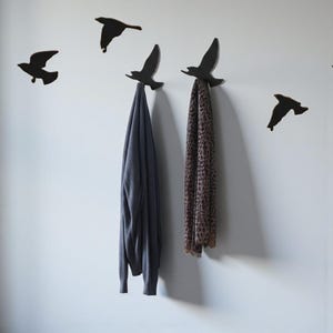 May include: Decorative wall hooks in the shape of flying birds. The hooks are black and hold a gray sweater and a brown and tan patterned scarf. The hooks are mounted on a white wall.