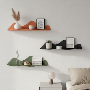 Puede incluir: Tres estanterías de pared modernas con forma de onda en naranja, negro y verde. Cada estantería contiene objetos decorativos como jarrones, pequeñas plantas, arte enmarcado y objetos decorativos. Las estanterías están montadas en una pared blanca.