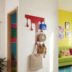May include: A red metal wall-mounted coat rack with a unique, dripping design. Several handbags and purses in various styles and colors hang from the hooks. The rack is mounted on a white wall, with a glimpse of a colorful interior.