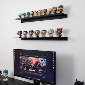 Puede incluir: Estanterías negras montadas en la pared que muestran una colección de figuras Funko Pop. Las estanterías están encima de un monitor de ordenador y un teclado. El monitor muestra una interfaz colorida. Las figuras son de varios personajes.