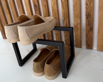 May include: Two pairs of beige suede loafers displayed on a black metal shoe rack. The shoes have a classic design with a rounded toe and a low profile sole. The rack has a minimalist, geometric design.