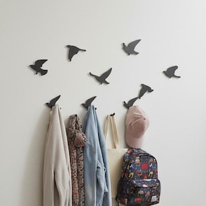 Op de afbeelding: Een witte muur met zwarte vogelvormige haken, met een lichtgekleurde jas, een sjaal met patroon, een spijkerjas en een draagtas. Een roze pet en een rugzak hangen ook. De haken zijn zo gerangschikt dat ze vogels in vlucht nabootsen.