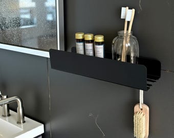 May include: A black metal shelf mounted on a dark gray wall, holding small bottles with gold lids, a glass jar with toothbrushes, and a wooden brush. The shelf is part of a bathroom setup.