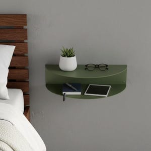 Könnte beinhalten: Ein grünes, halbkreisförmiges Wandregal mit einer kleinen weißen Topfpflanze, einer Brille, einem Notizbuch, einem Stift und einem Tablet. Das Regal ist an einer grauen Wand befestigt, mit einem hölzernen Kopfteil und Bett links.