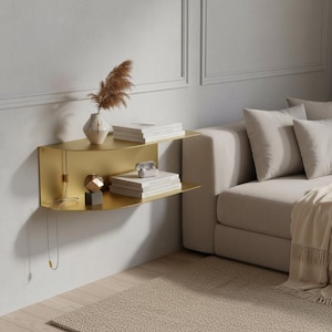 Puede incluir: Una estantería dorada montada en la pared con dos niveles. La estantería superior contiene libros y un jarrón con flores secas. La estantería inferior muestra libros y objetos decorativos. La estantería está montada en una pared blanca junto a un sofá beige.