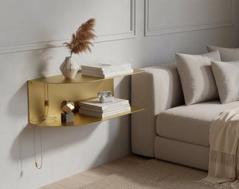 May include: A gold-colored, wall-mounted shelf with two tiers. The top shelf holds books and a vase with dried flowers. The bottom shelf displays books and decorative objects. The shelf is mounted on a white wall next to a beige sofa.