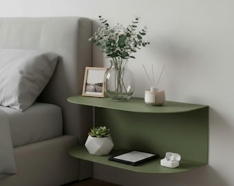May include: A modern, olive green wall-mounted nightstand with two shelves. The top shelf holds a clear glass vase with flowers, a framed photo, and a diffuser. The bottom shelf displays a succulent in a white geometric pot, an e-reader, and a pair of white wireless earbuds.