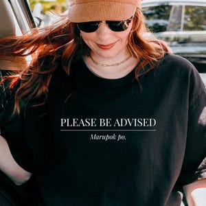 Puede incluir: Camiseta negra con el texto "PLEASE BE ADVISED" y "Marupok po." en blanco. La persona de la imagen lleva un sombrero de pana marrón y gafas de sol. La persona está sentada en un coche.