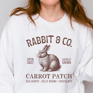 Puede incluir: Sudadera blanca con un gráfico de estilo vintage. El diseño presenta el texto "RABBIT & CO." encima de una ilustración detallada de un conejo. Debajo del conejo, se muestra el texto "CARROT PATCH", junto con los textos "OPEN DAILY", "LOCALLY GROWN", "EGG HUNTS", "JELLY BEANS" y "CHOCOLATE".