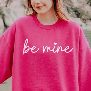 May include: A pink sweatshirt with the text "be mine" in white cursive lettering with a small heart above the "e".
