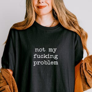May include: Black t-shirt with the text "not my fucking problem" in white. The shirt is paired with a brown jacket. The image suggests a casual, relaxed style, with the text as a statement.